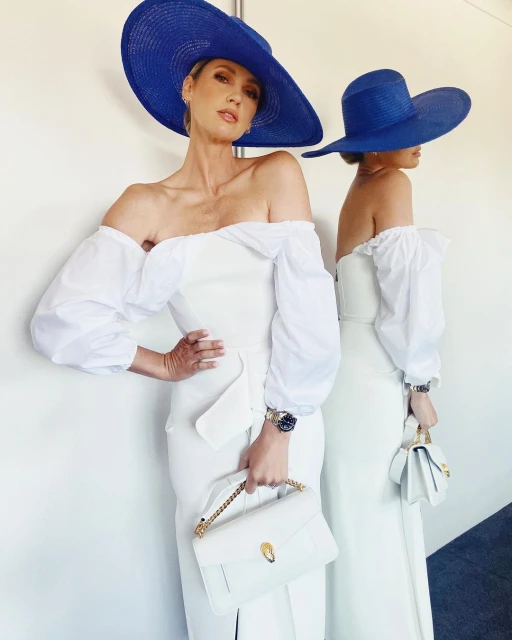Woman wearing a large blue hat and off-the-shoulder white dress holding a white handbag, with a mirror reflection behind her.