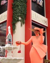Woman in bright orange dress and wide-brim hat sabering a champagne bottle by a stone fountain in Sydney