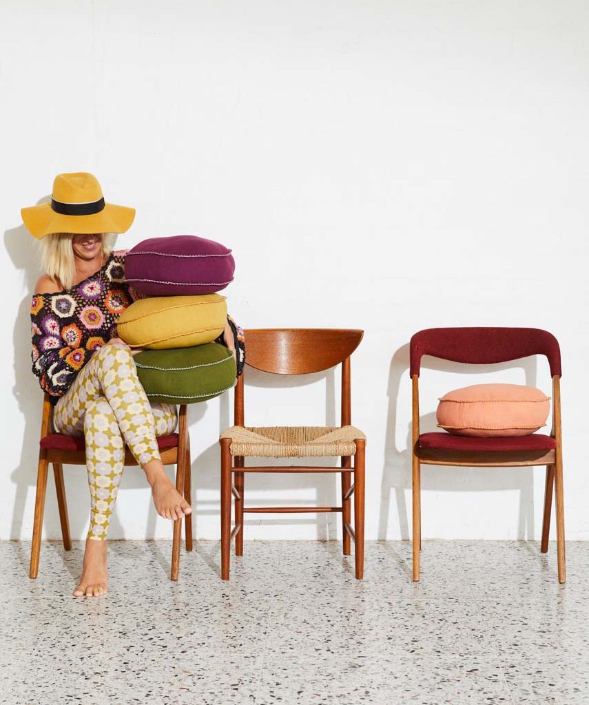 Blonde woman in floral sweater and yellow hat poses barefoot sitting with colorful cushions stacked on lap next to chairs.