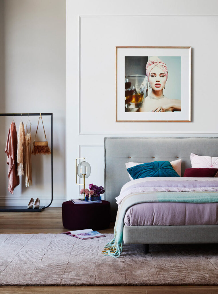 Bedroom interior featuring a grey upholstered bed, framed portrait art, a clothing rack, and lilac-toned décor details.