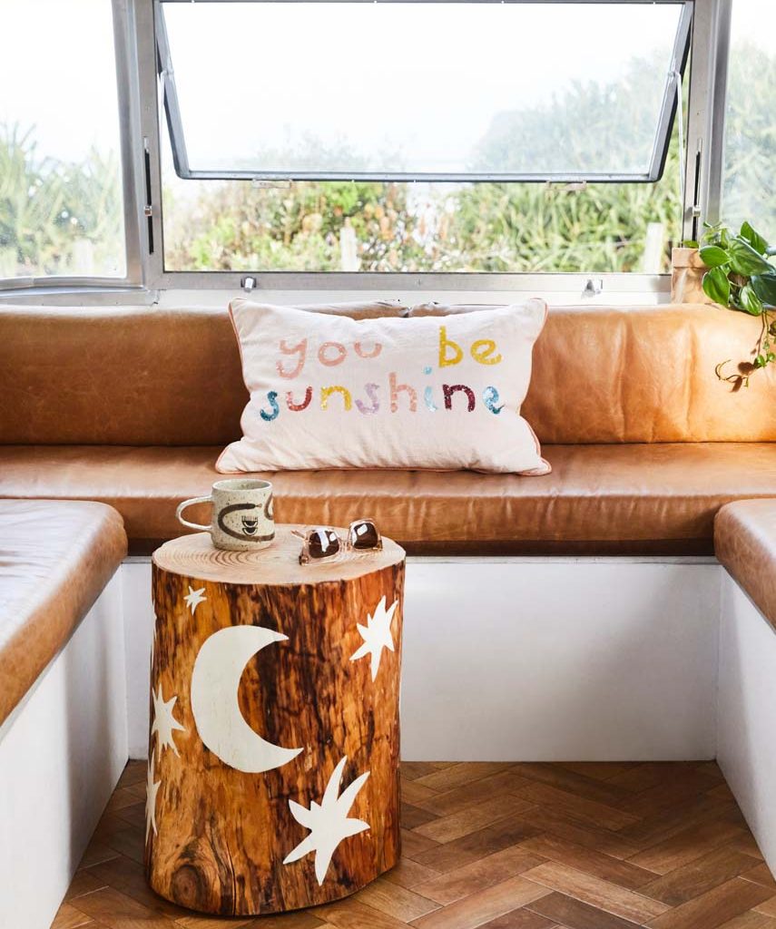 Interior view of a camper featuring a wooden stump table with moon and star designs, a pillow with text, and a cozy seating area.