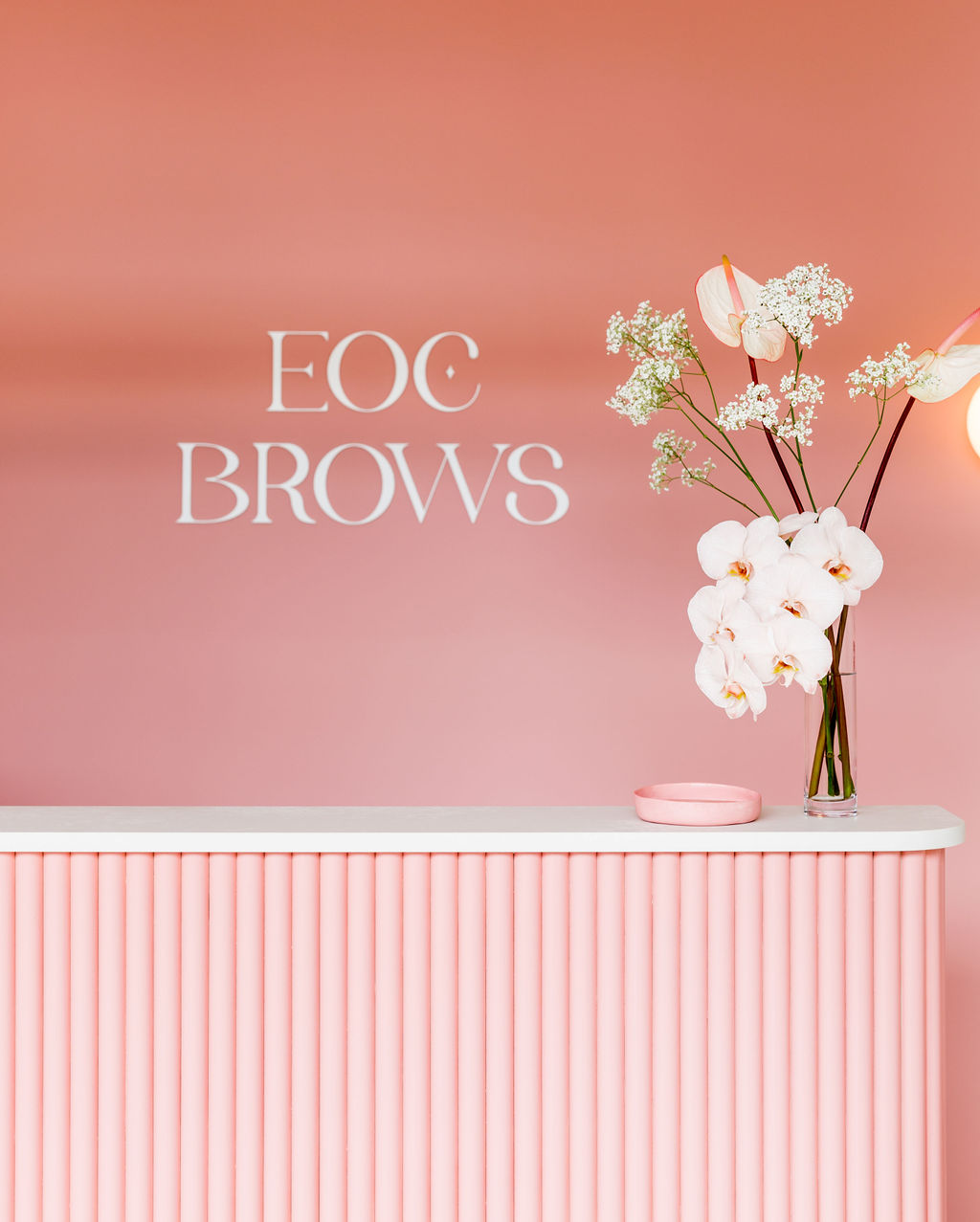 Reception area featuring a pink counter with vertical tubes, white orchids in a glass vase, and a pink wall with 'EOC Brows' text.