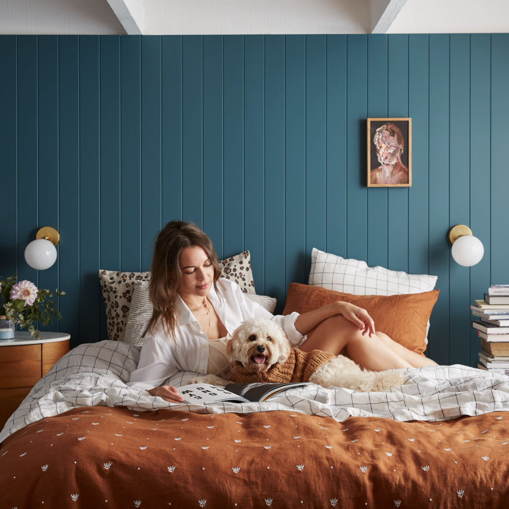 Woman and dog relax on a bed with blue paneled wall, throw pillows, white sheets, rust colored coverlet, and bedside table with flowers.