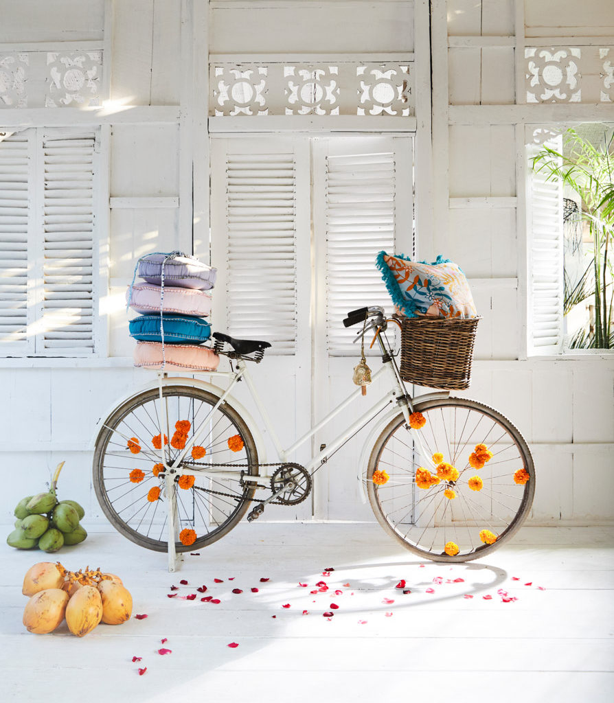 Vintage white bicycle with floral accents, carrying pillows in a basket and on the seat, stands against a white shuttered wall.