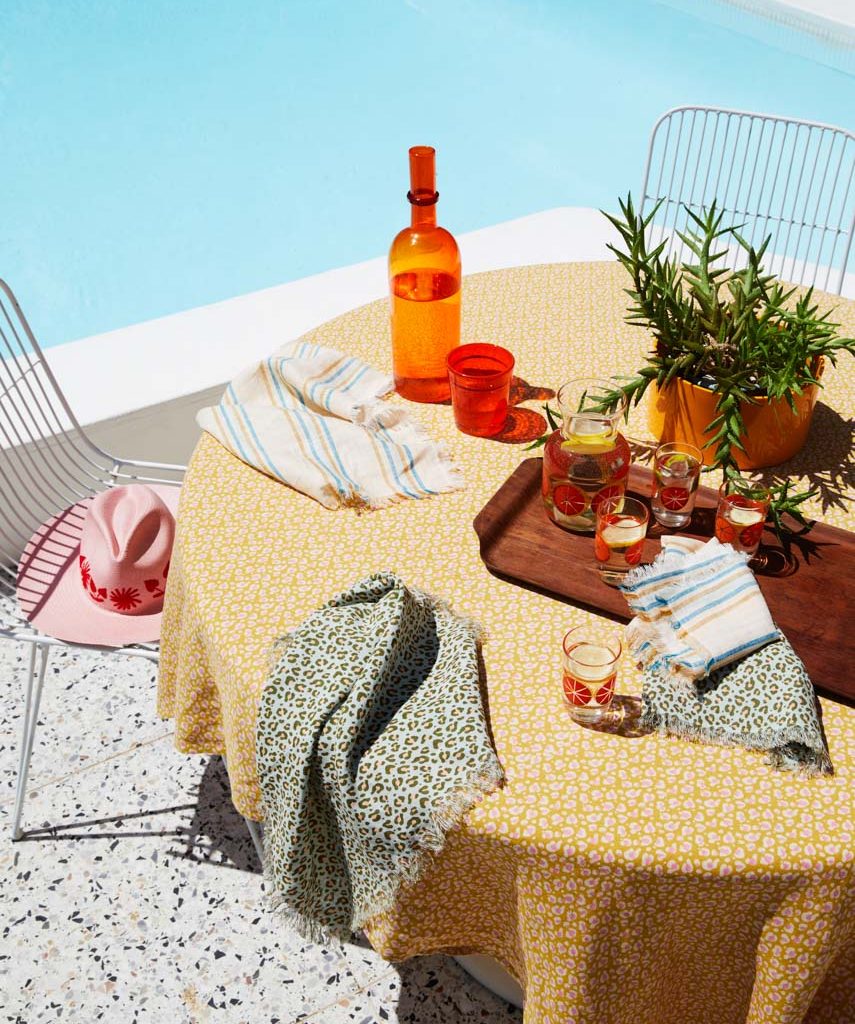 Outdoor table setting with a yellow floral tablecloth, orange glassware, linens, a wooden tray, and a potted plant by the pool.