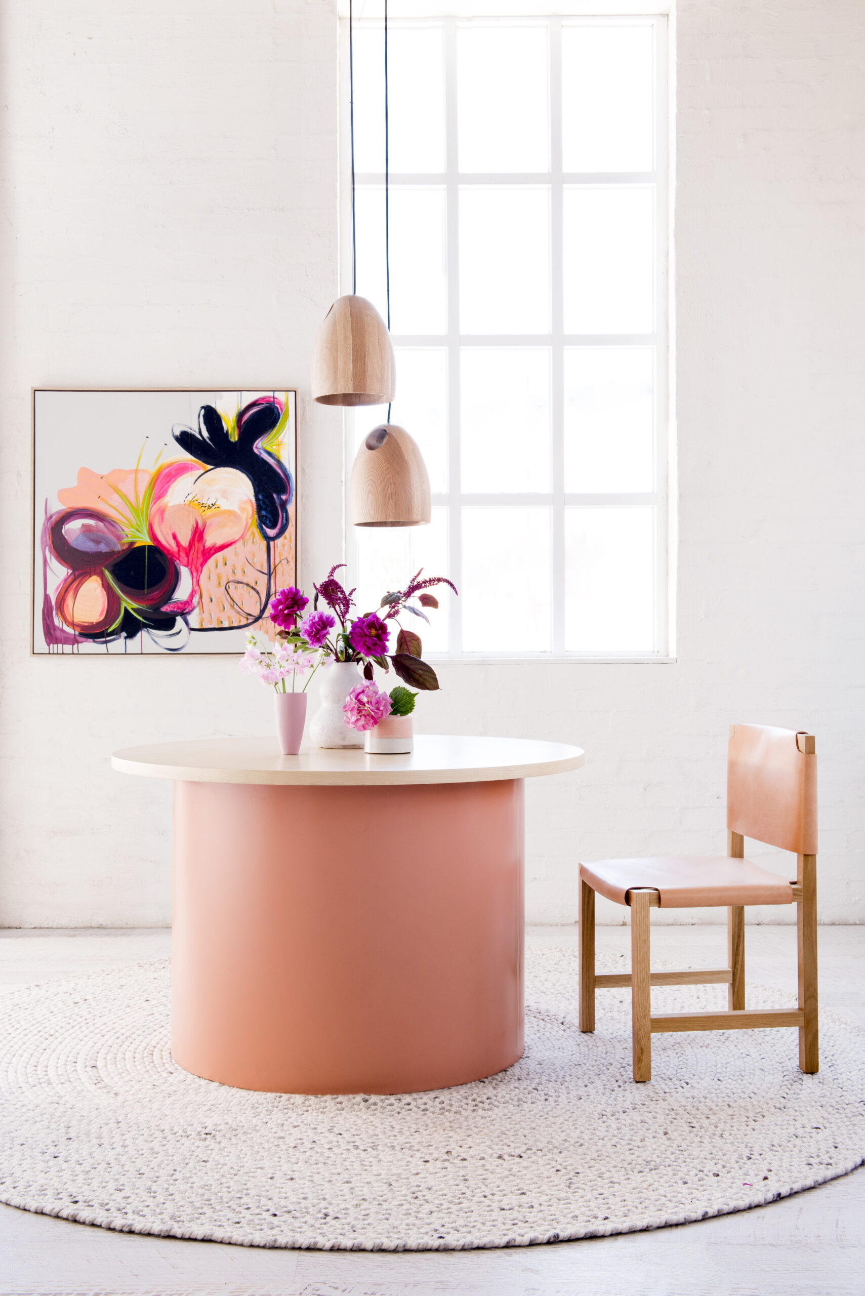Interior photograph of a round table with flowers, wooden chair, and pendant lights in a bright white room with a modern art piece.
