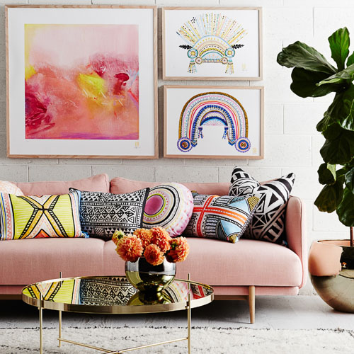 Living room setup with a pink couch adorned with patterned throw pillows, a reflective coffee table, vibrant artwork, and an indoor plant.