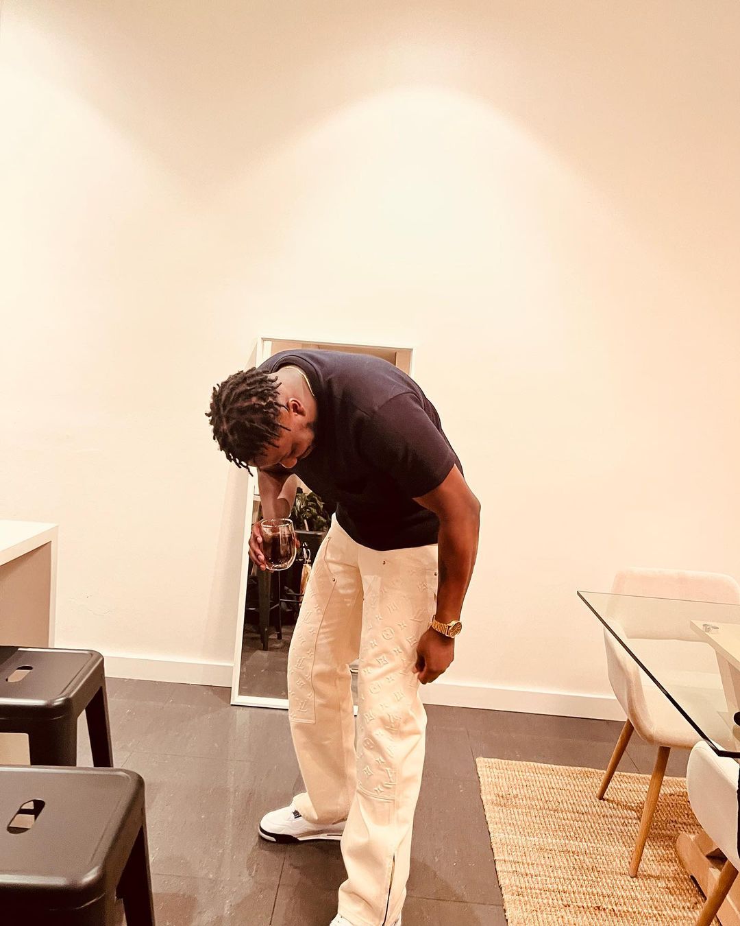 Adult male with dreadlocks in black t-shirt and light patterned pants holding a glass indoors, near a mirror and modern dining table.