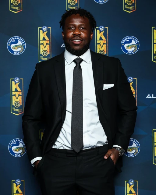 Man in suit and tie posing on a branded event backdrop with team logos in a studio shot with eye-level angle, promoting sports and entertainment.