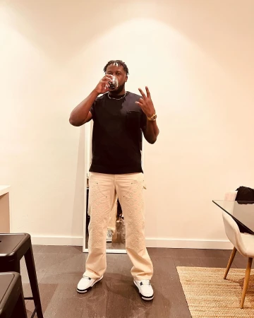 Black man drinking from a glass and flashing a peace sign while wearing a black t-shirt and cream patterned trousers