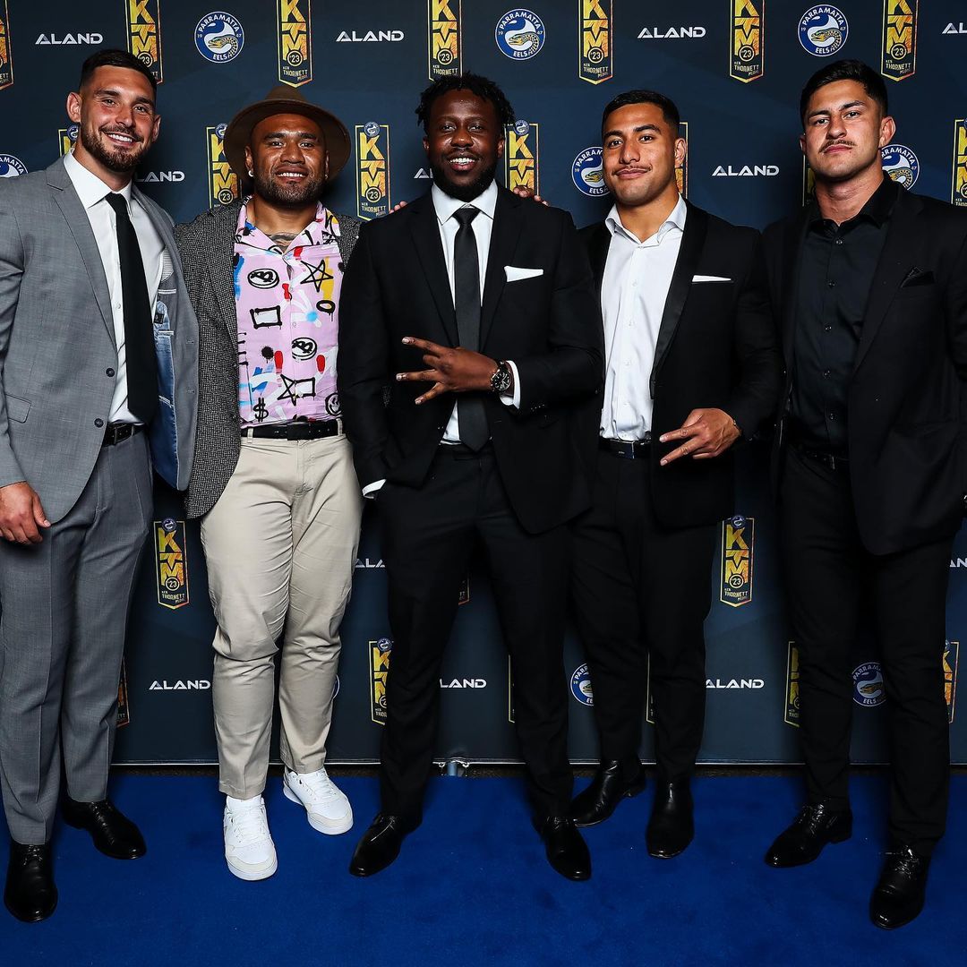 Five men in formal wear pose against a branded backdrop. The group includes a mix of fashion choices, all standing on a blue carpet.