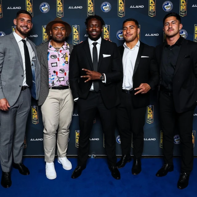 Five men in formal wear pose against a branded backdrop. The group includes a mix of fashion choices, all standing on a blue carpet.