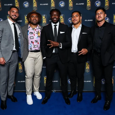 Five men in formal wear pose against a branded backdrop. The group includes a mix of fashion choices, all standing on a blue carpet.
