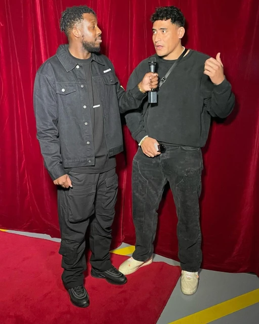 Two men in black clothing are talking on a red carpet in front of a red curtain, one holding a microphone, in an indoor setting.