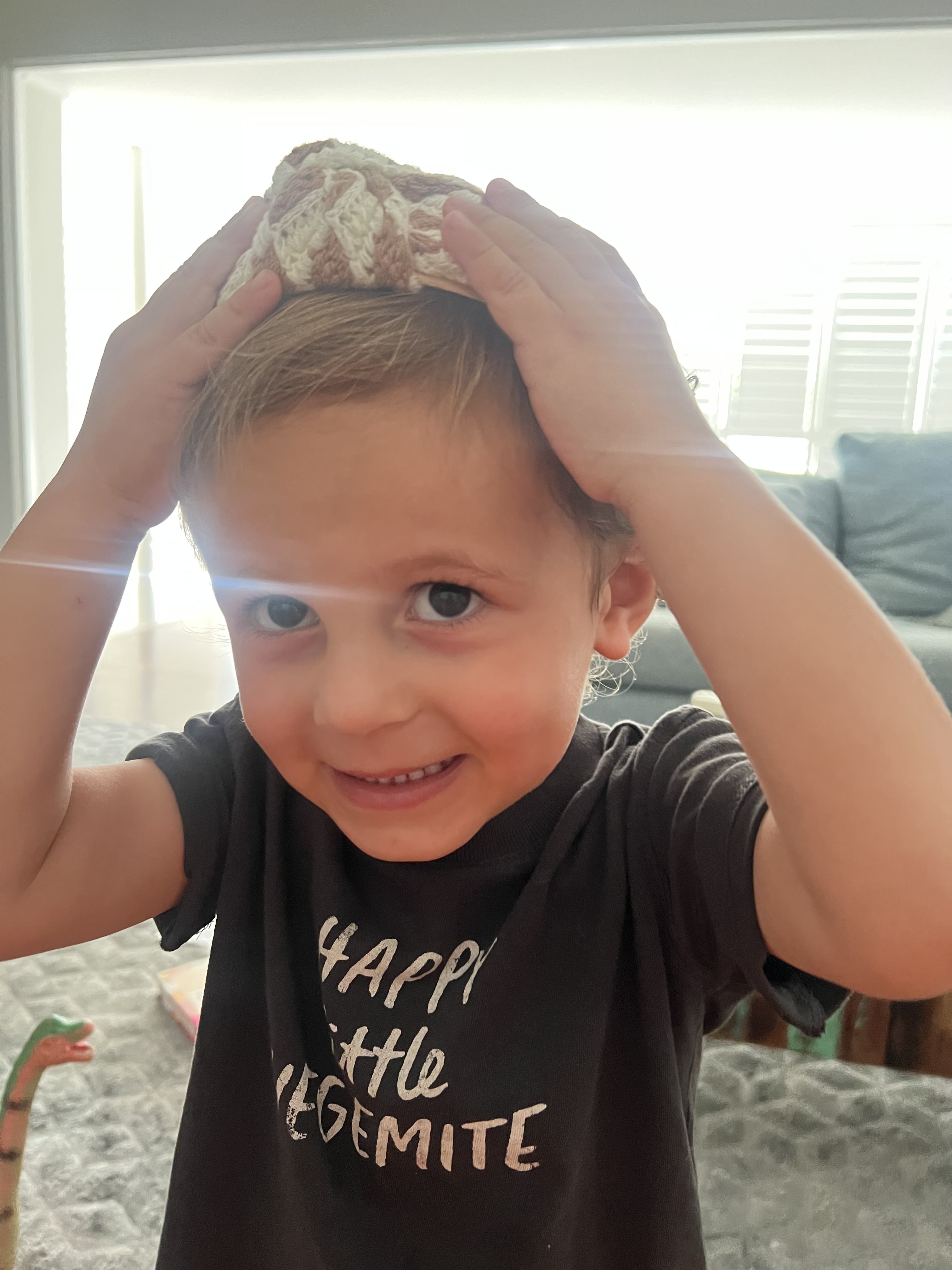 Smiling young child wearing a crocheted hat while indoors, looking directly at the camera, with window light in the background.