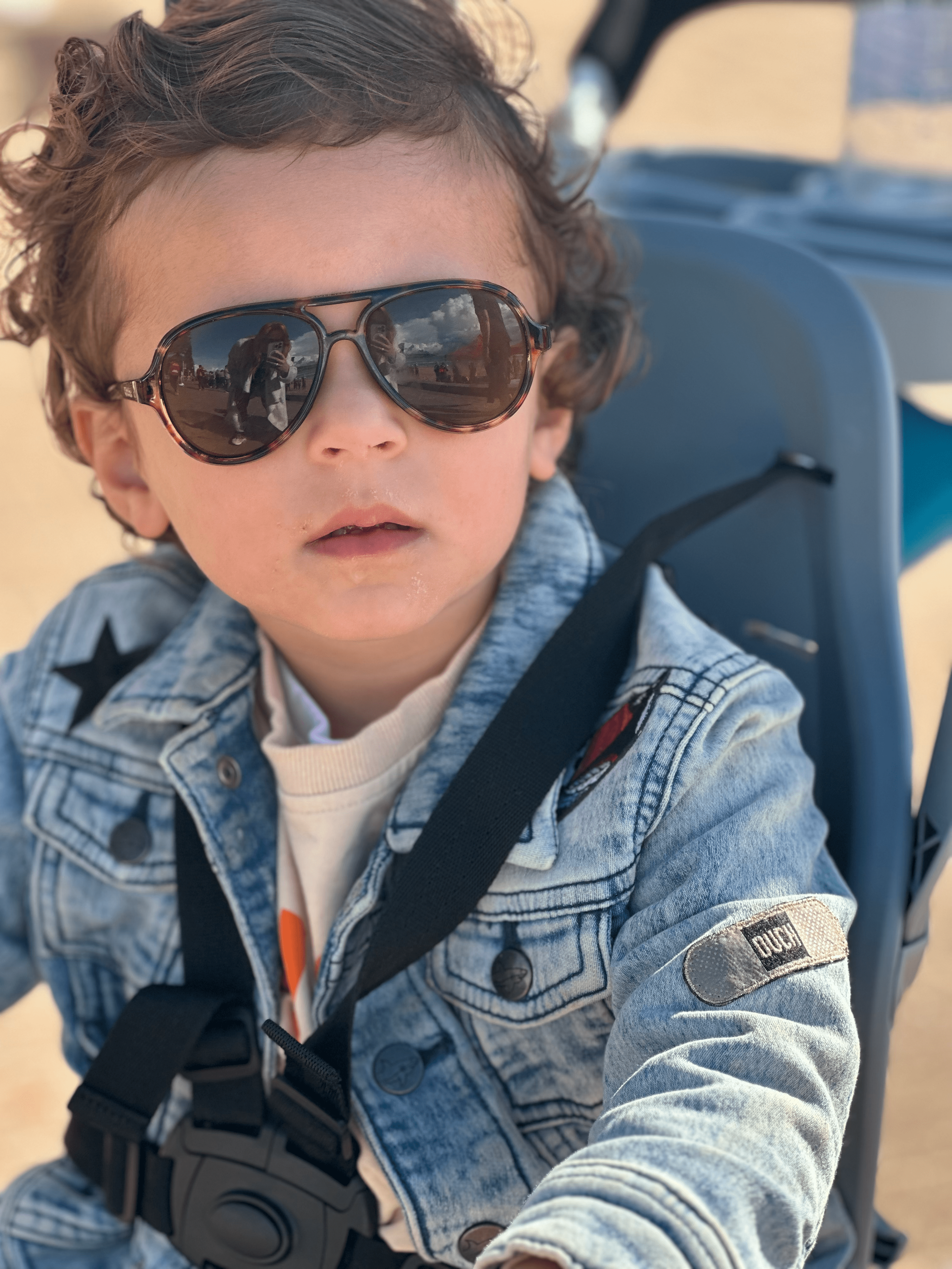 Toddler wearing sunglasses and a denim jacket sits in a stroller, enjoying the day outdoors with a blurry outdoor background.
