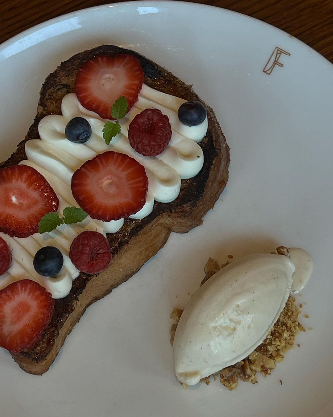 Overhead shot of a slice of French toast topped with whipped cream and berries, alongside a quenelle of dessert on a white plate.