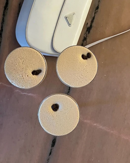 Three espresso martini cocktails garnished with coffee beans are displayed with a white purse on a pink marble surface.
