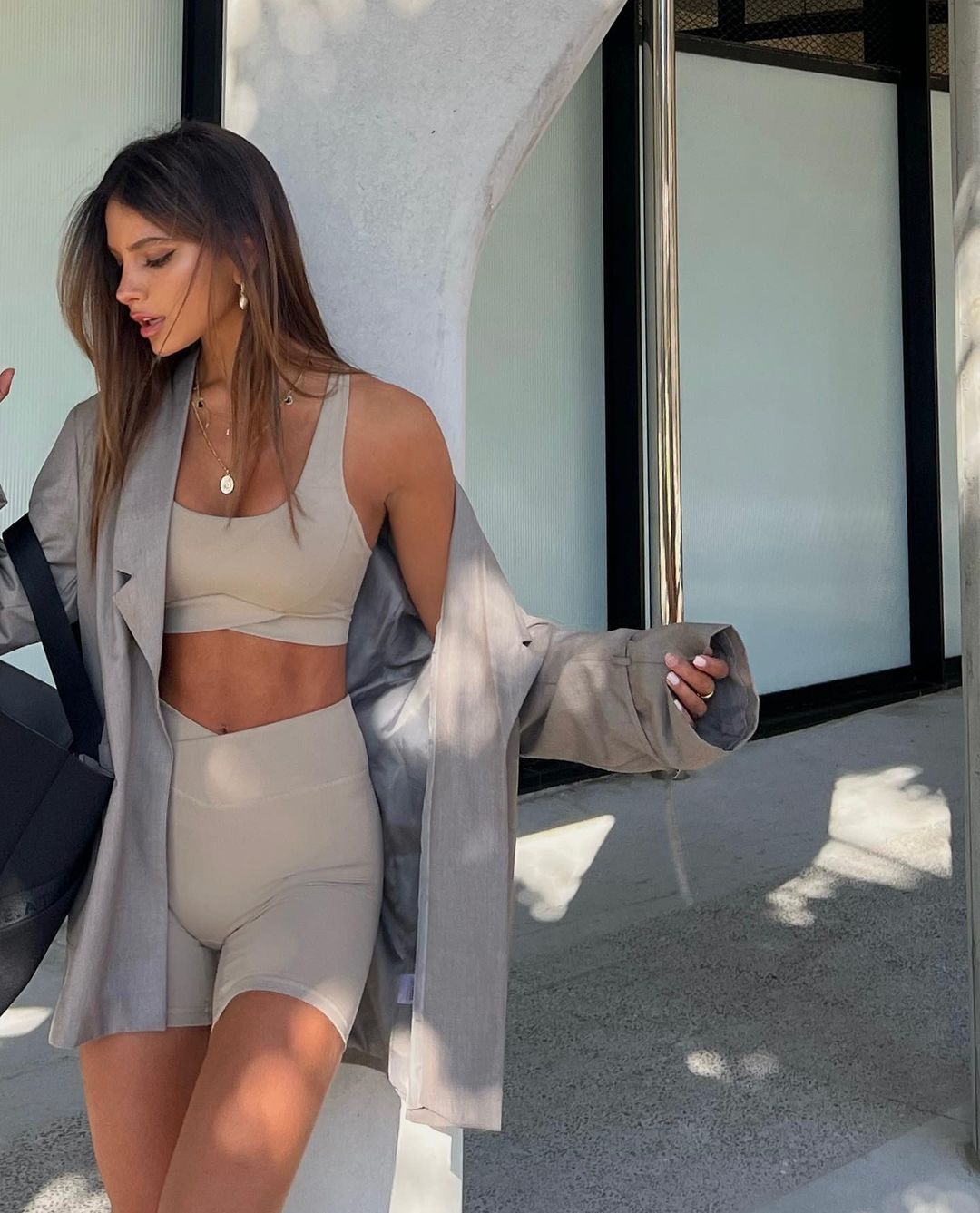 Attractive woman in a beige two-piece activewear set and oversized grey blazer poses near a textured wall and black pillar.