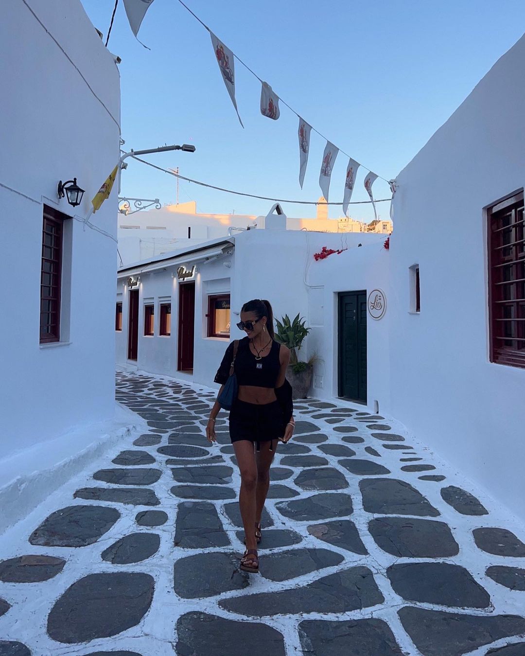 Woman in black crop top and skirt walking on cobblestone street lined with white buildings and hanging flags on a sunny day.