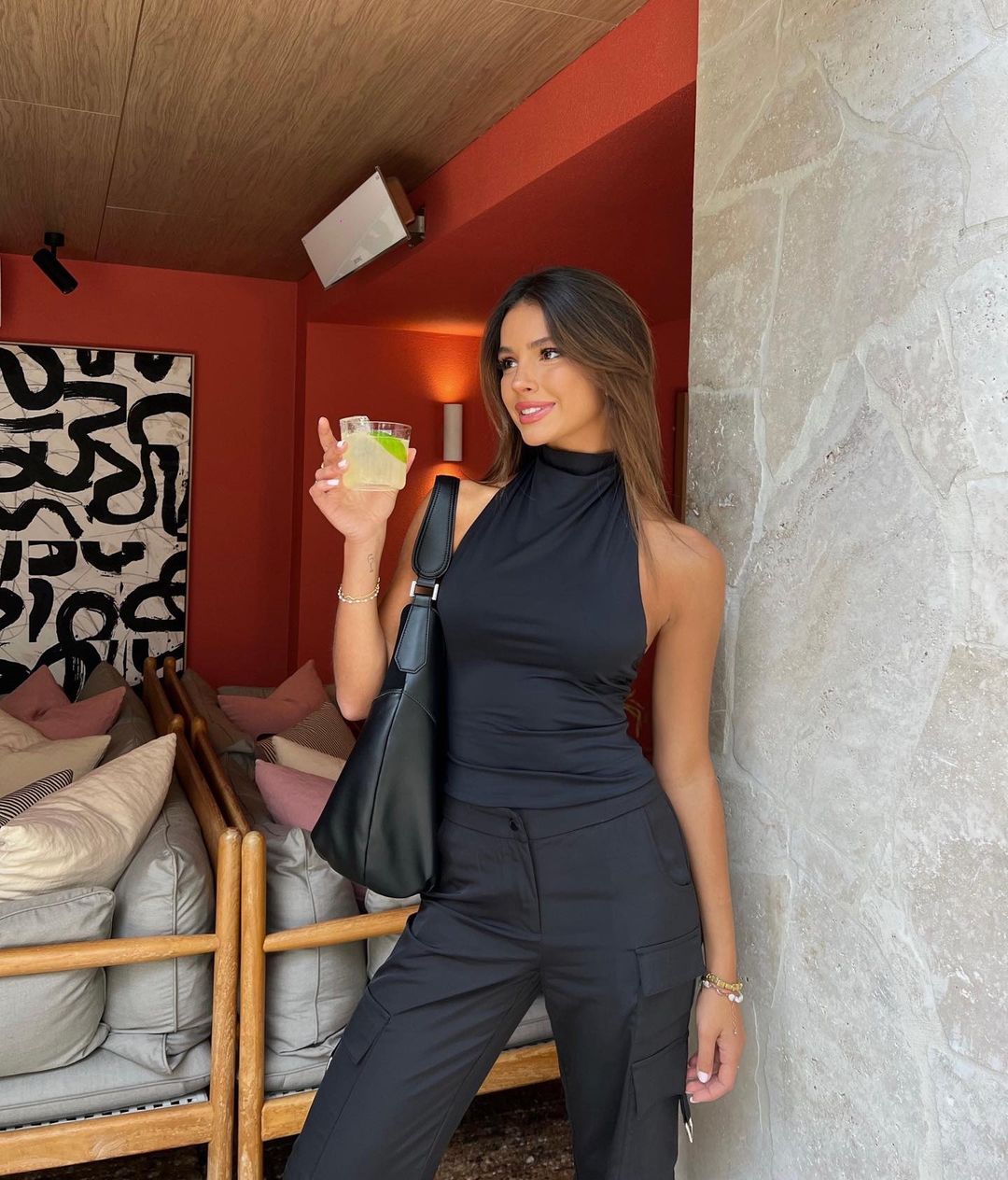 A stylish woman in a black outfit smiles while holding a cocktail in an indoor setting with modern decor and soft lighting.