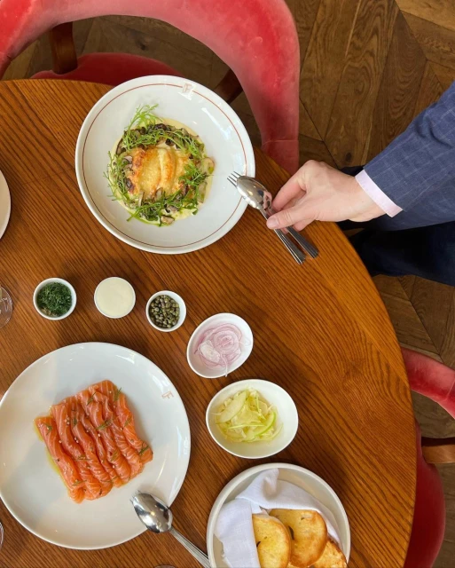 Overhead shot of a wood table setting with fish and cheese appetizers, small bowls of condiments, toast, and a suited person's hand holding silverware.