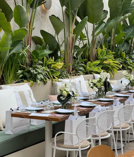 Outdoor banquet setup with long wooden table, white chairs, floral arrangements, and lush greenery in the background, daytime.