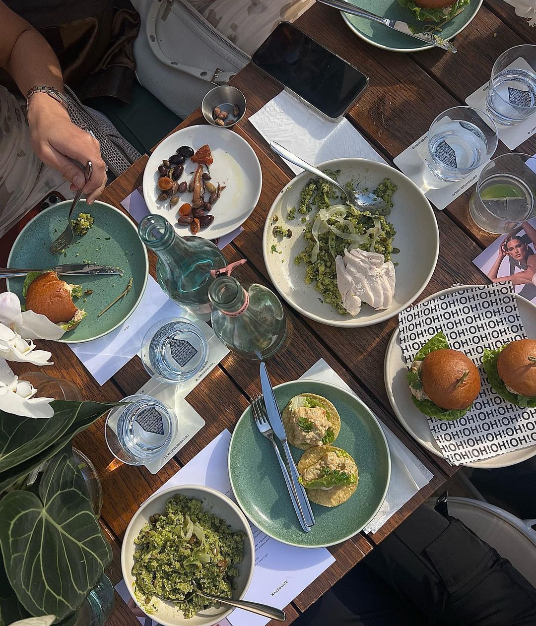 Overhead shot of a wooden table setting with various plates of food, drinks, and dining utensils. A smartphone sits on the table.