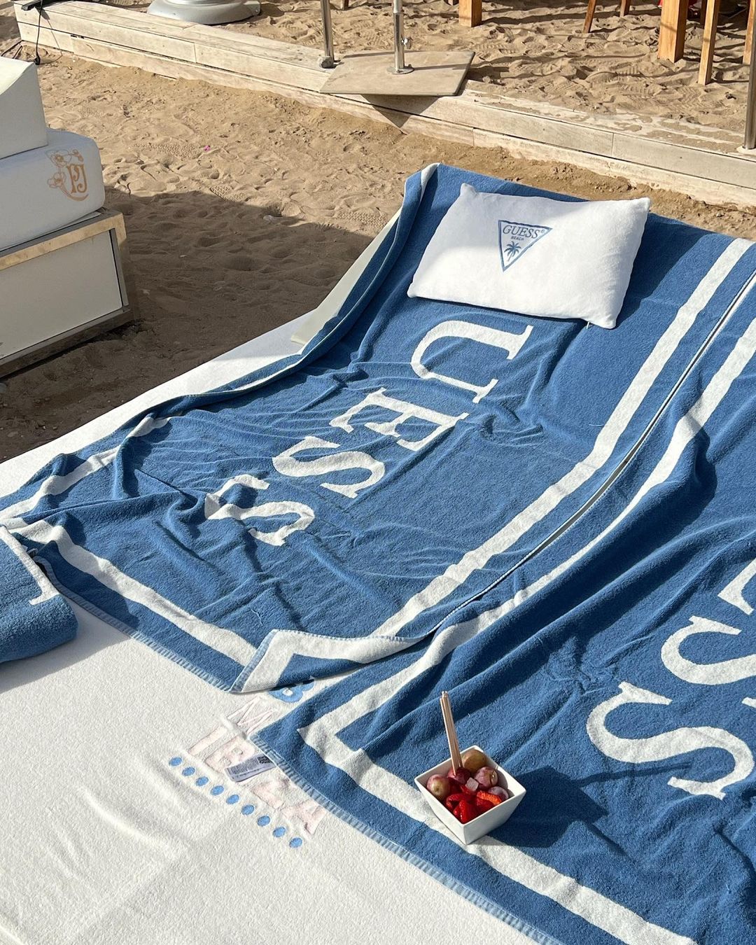 Outdoor lounge setup on a beach with a blue and white striped towel, white pillow, fruit bowl, and beach furniture.