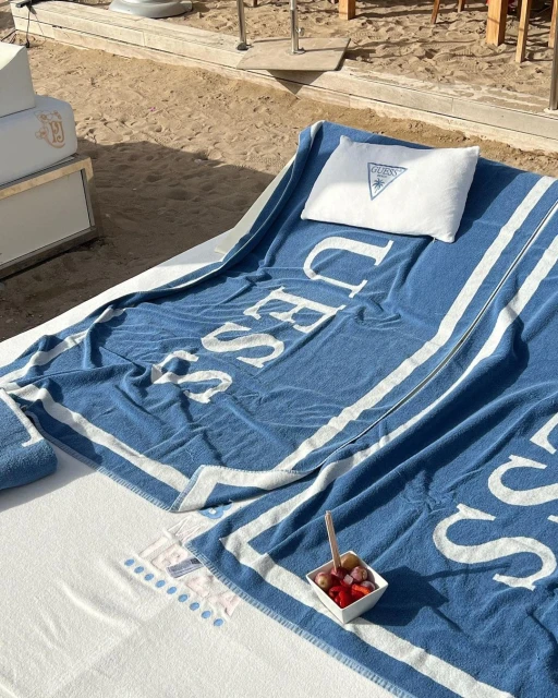 Outdoor lounge setup on a beach with a blue and white striped towel, white pillow, fruit bowl, and beach furniture.