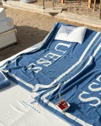 Blue beach towel with white lettering spread across a white lounge sunbed on sand