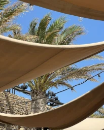 Palm tree and string lights viewed beneath layered beige fabric shade sails against a clear blue sky.