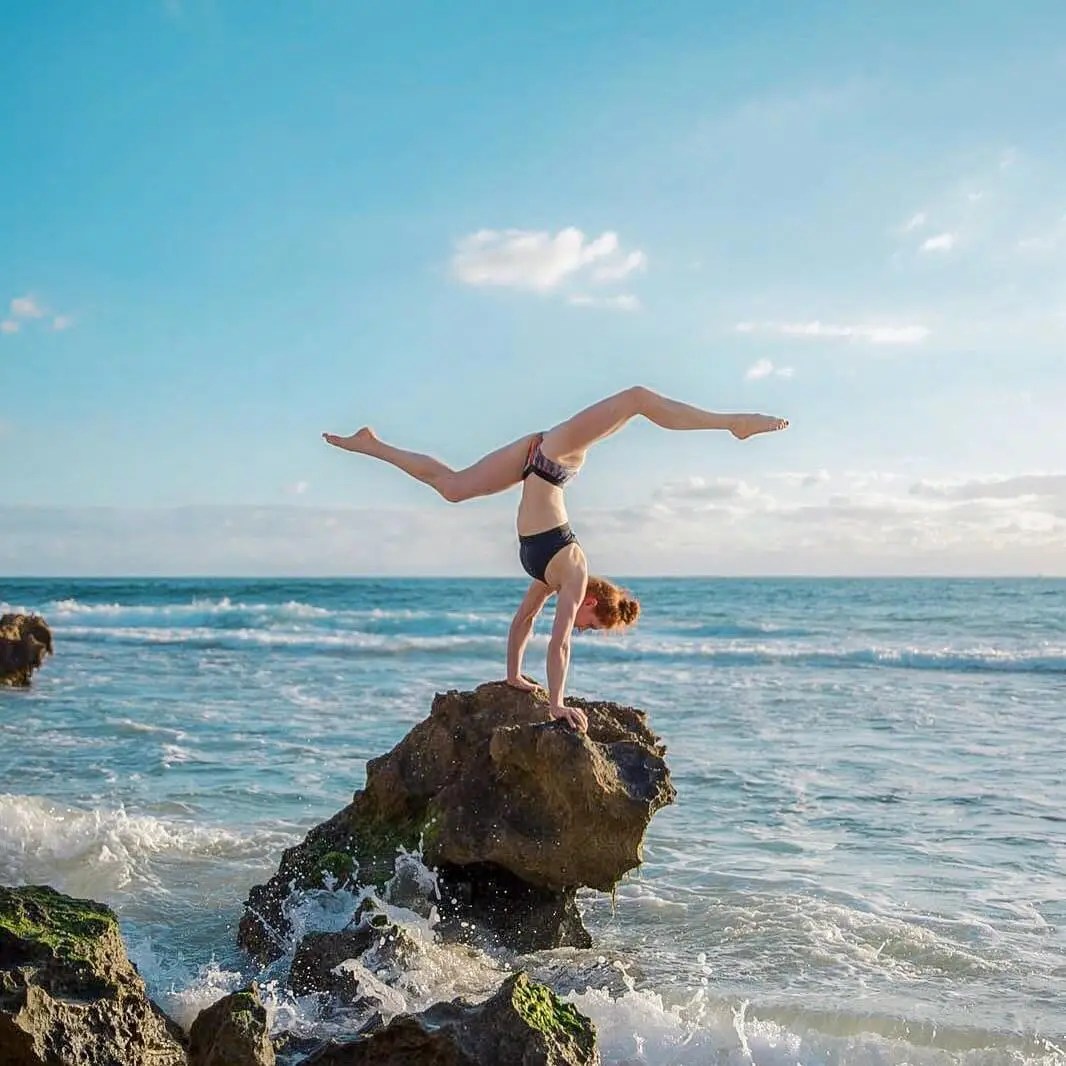Athletic woman in a bikini performing a handstand on ocean rocks. She is illuminated by natural light and has her legs spread wide.
