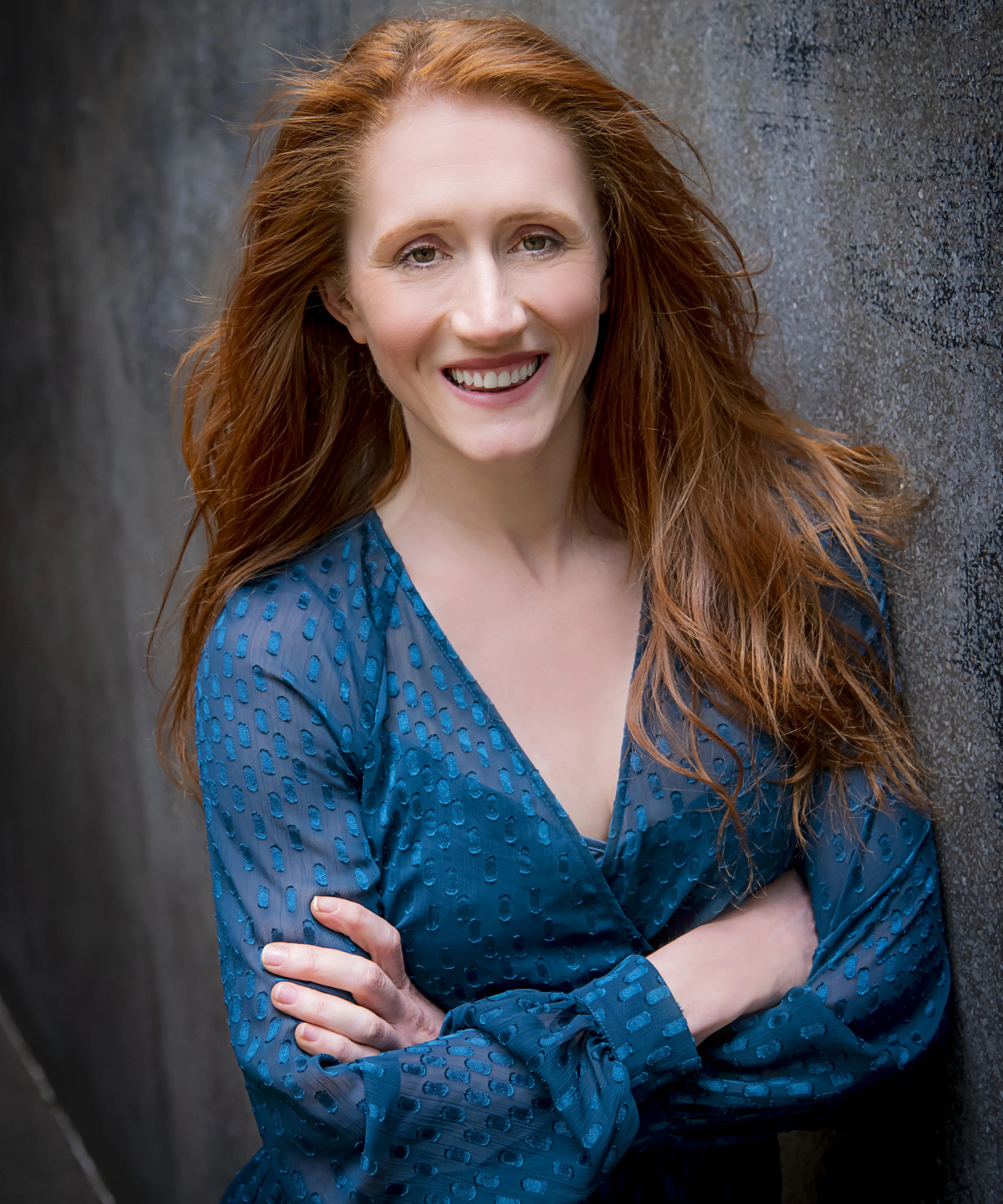Close-up portrait of an adult woman with long red hair, smiling and wearing a blue patterned top, posing in front of a textured gray background.