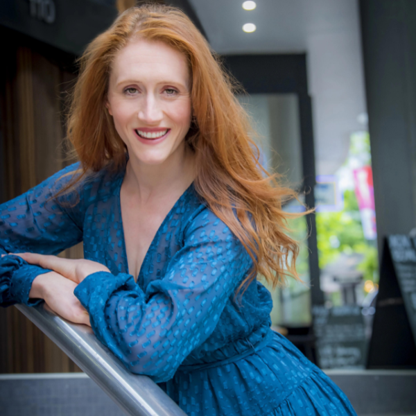 Portrait of a smiling, red-haired woman in a blue dress leaning against a railing outdoors with a building visible in the background.