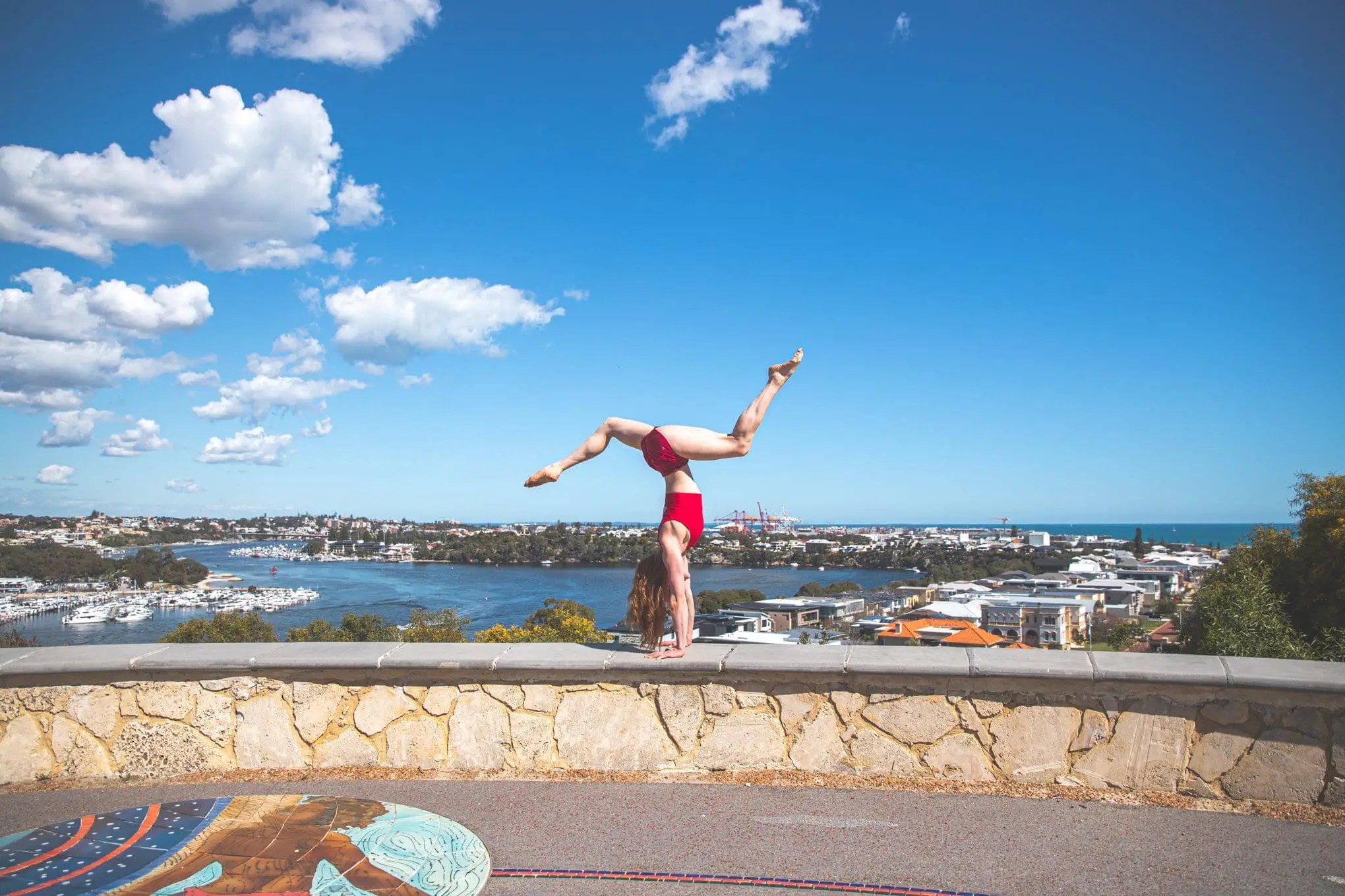 Adult female does a handstand on a stone wall overlooking a city and harbor under a sunny sky with scattered white clouds.