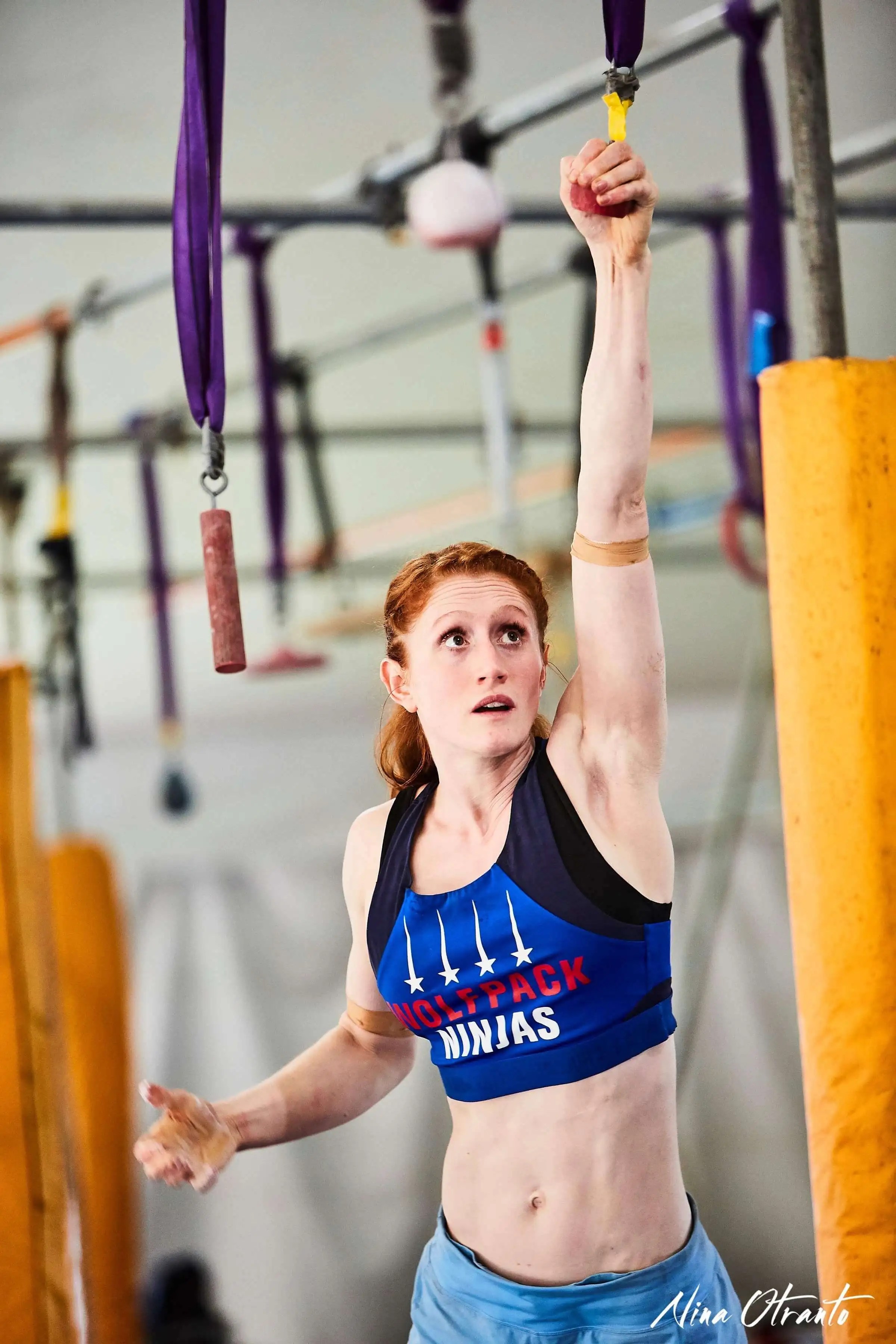 Female athlete with red hair in sports bra hangs from a yellow bar at a ninja warrior obstacle course, showcasing strength and athleticism.