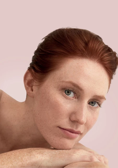 Close-up portrait of a light-skinned adult female with freckles and auburn hair in a beauty or skincare context against a pale pink backdrop.