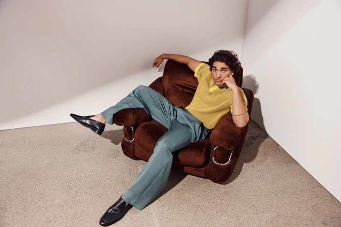 A man reclines in a brown armchair, wearing a yellow shirt and blue-green pants, creating a relaxed fashion-forward portrait.