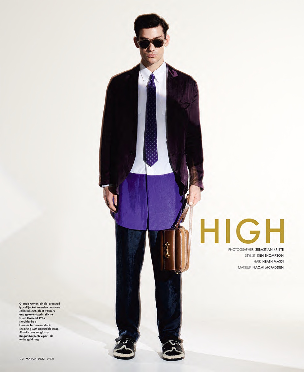 Male model poses in a purple blazer, white shirt, navy tie, blue pants, and black shoes against a bright white studio backdrop.