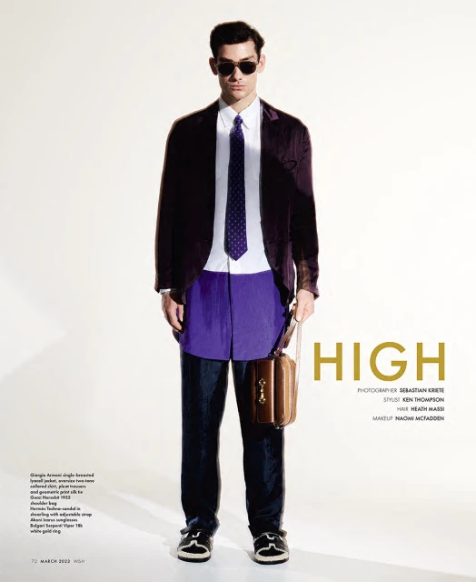 Male model poses in a purple blazer, white shirt, navy tie, blue pants, and black shoes against a bright white studio backdrop.