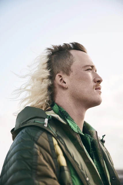 Portrait of a man with a half-blonde mohawk hairstyle wearing a green jacket, looking up towards the sky in an outdoor setting.