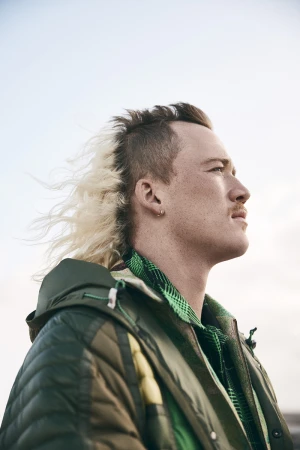 Portrait of a man with a half-blonde mohawk hairstyle wearing a green jacket, looking up towards the sky in an outdoor setting.