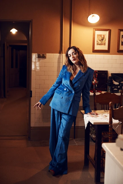 A woman poses indoors, wearing a blue velvet double-breasted suit and leaning against a table with a white tablecloth in an interior setting.