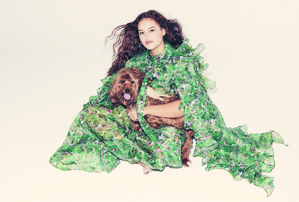 A young woman in a flowing green floral dress hugs a fluffy brown dog in a studio shot against a light background.