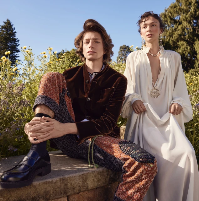 Two fashion models pose in garden. Man in brown jacket and patterned pants sits on stone. Woman in white dress stands beside him.