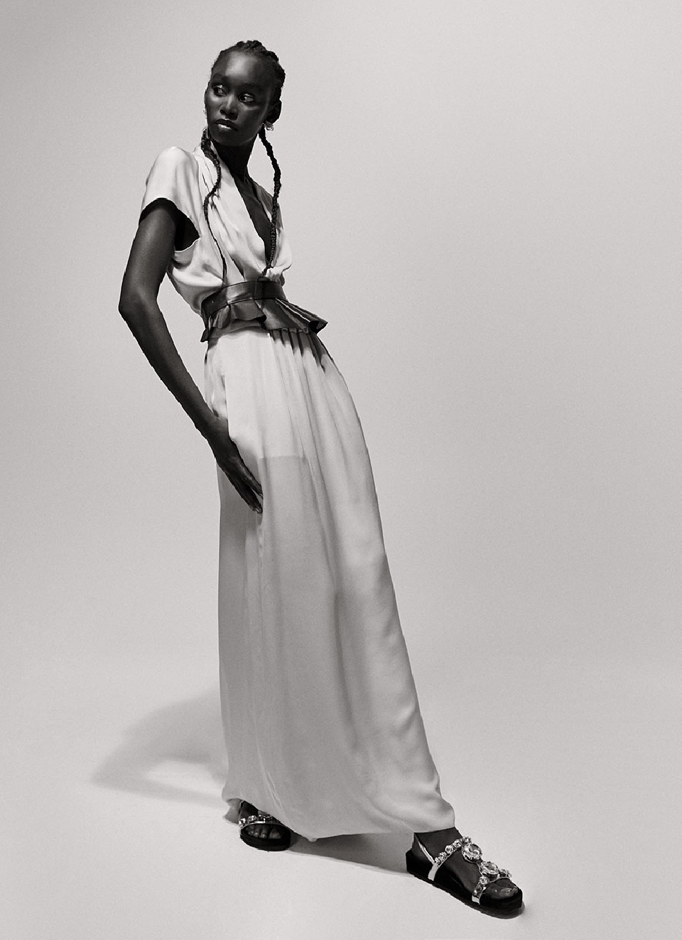 Full-length portrait of an African-American fashion model wearing a light-colored dress with a pleated belt and embellished sandals in a studio.