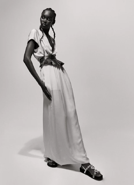 Full-length portrait of an African-American fashion model wearing a light-colored dress with a pleated belt and embellished sandals in a studio.