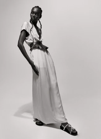 Full-length portrait of an African-American fashion model wearing a light-colored dress with a pleated belt and embellished sandals in a studio.