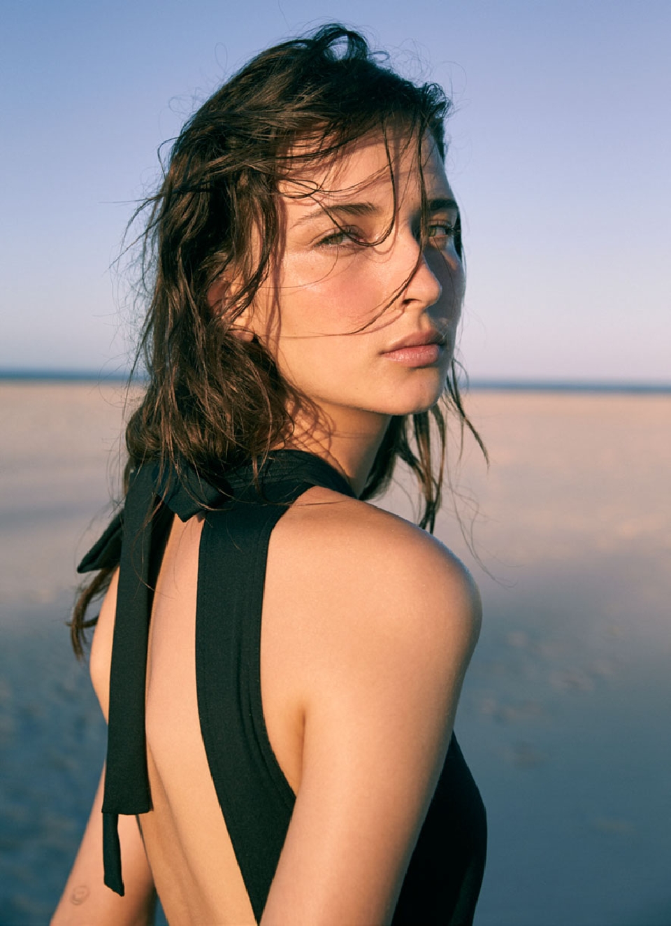 Adult female portrait on beach with a black dress and wet hair, blue sky in the background, captured with natural lighting.